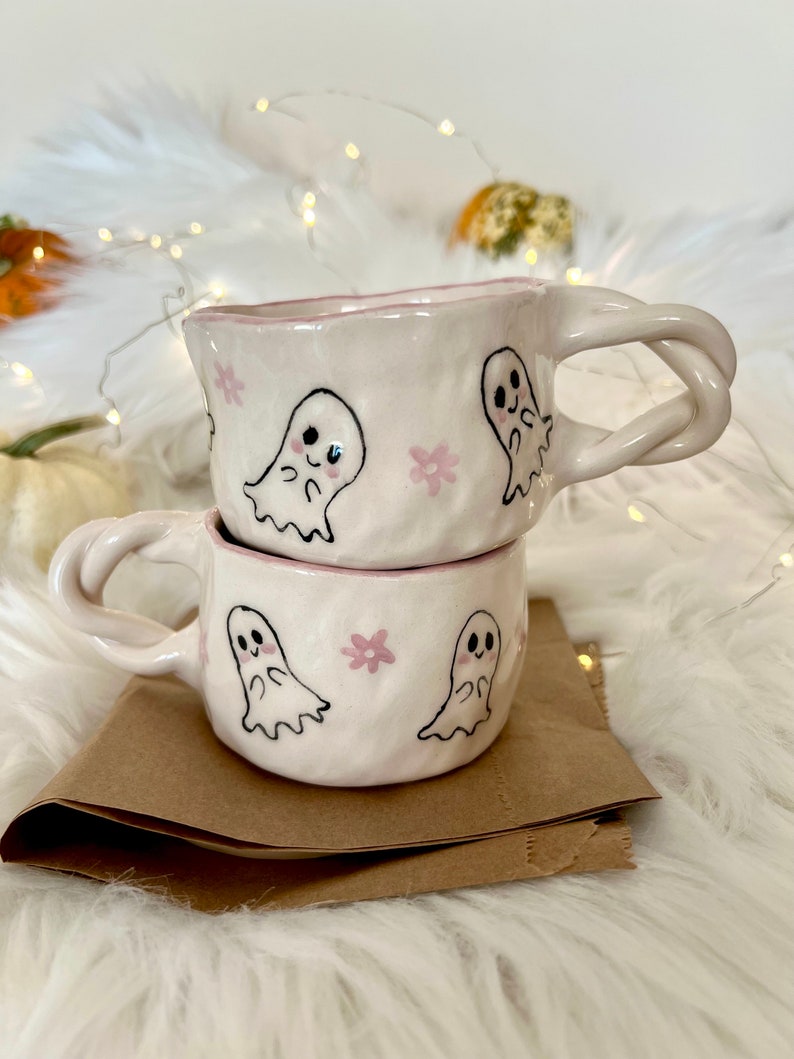 May include: Two white ceramic mugs with pink floral and ghost designs. The mugs are stacked on top of each other. The mug on top has a twisted handle.