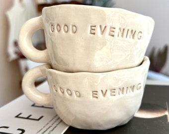 May include: Two white ceramic mugs stacked on top of each other. Both mugs have the words "Good Evening" embossed on the side.