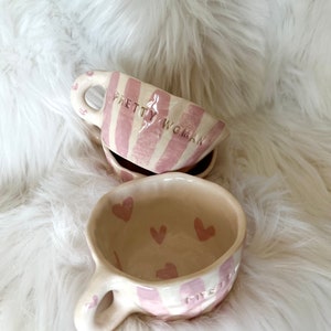 Puede incluir: Tazas de cerámica hechas a mano con rayas rosas y blancas y diseños de corazones. Una taza tiene las palabras "PRETTY WOMAN" y otra la palabra "MYSTERY" en el lateral. Las tazas tienen un asa con un diseño de corazón.