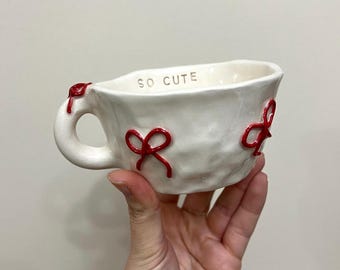 May include: A white ceramic mug with an irregular, hand-molded texture. The exterior features raised, glossy red bow decorations. The interior rim is inscribed with the text SO CUTE in dark lettering. The mug has a single handle with a red bow.