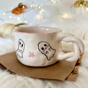May include: A white ceramic mug with a pink rim and a twisted handle. The mug is decorated with two cartoon ghosts and pink flowers.