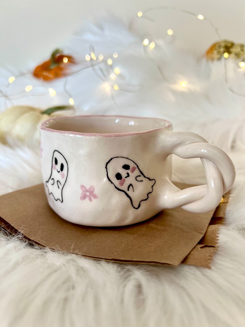 May include: A white ceramic mug with a pink rim and a twisted handle. The mug is decorated with two cartoon ghosts and pink flowers.