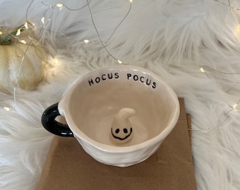 May include: White ceramic mug with a black handle. The mug has the words "Hocus Pocus" written on the side and a small ghost figurine inside. The mug is sitting on a brown paper bag.
