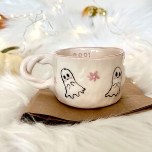 May include: A white ceramic mug with a pink rim and a pink and black ghost design. The mug says "BOO!" on the side.