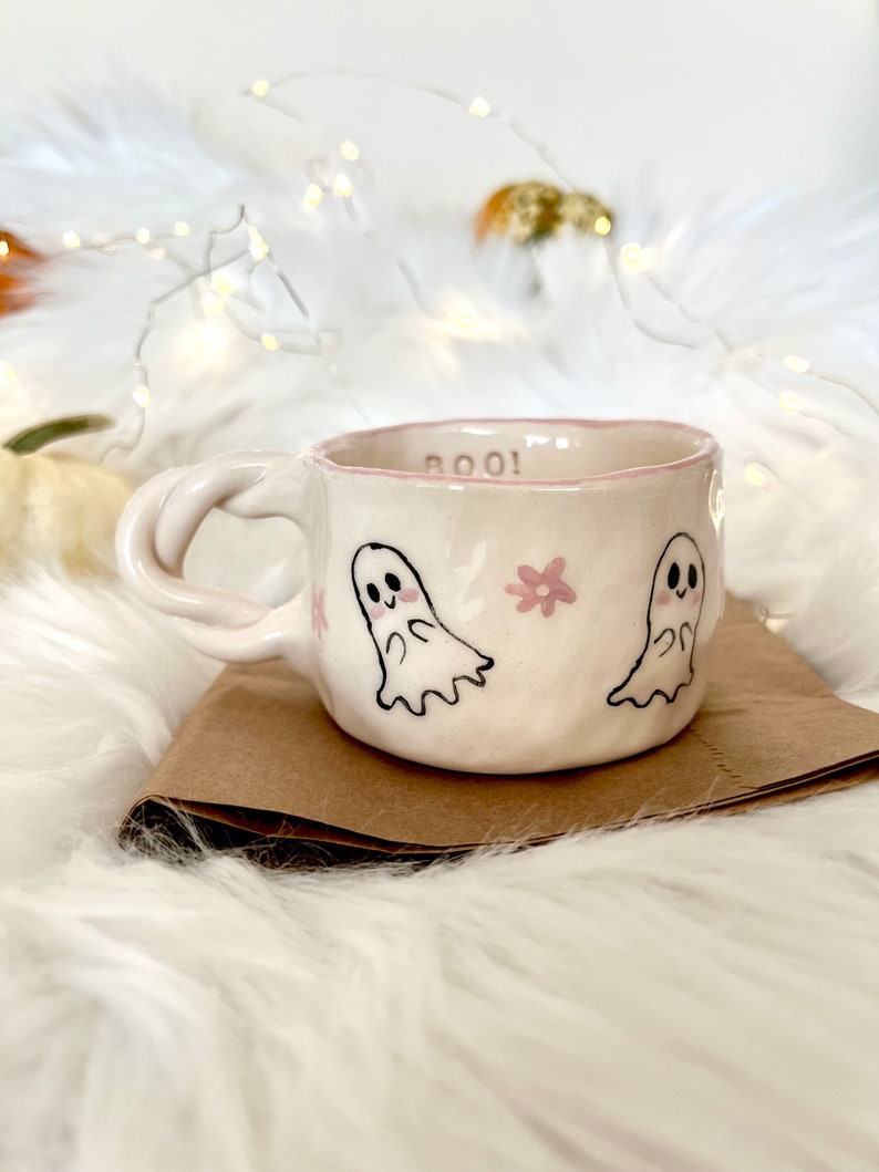May include: A white ceramic mug with a pink rim and a pink and black ghost design. The mug says "BOO!" on the side.