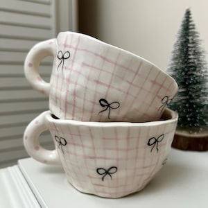 Puede incluir: Dos tazas de cerámica rosa y blanca con diseños de lazos negros. Las tazas están apiladas una encima de la otra. Las tazas tienen un patrón a cuadros.