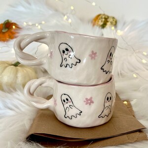 May include: Two white ceramic mugs with pink and black ghost illustrations and pink flowers. The mugs are stacked on top of each other.