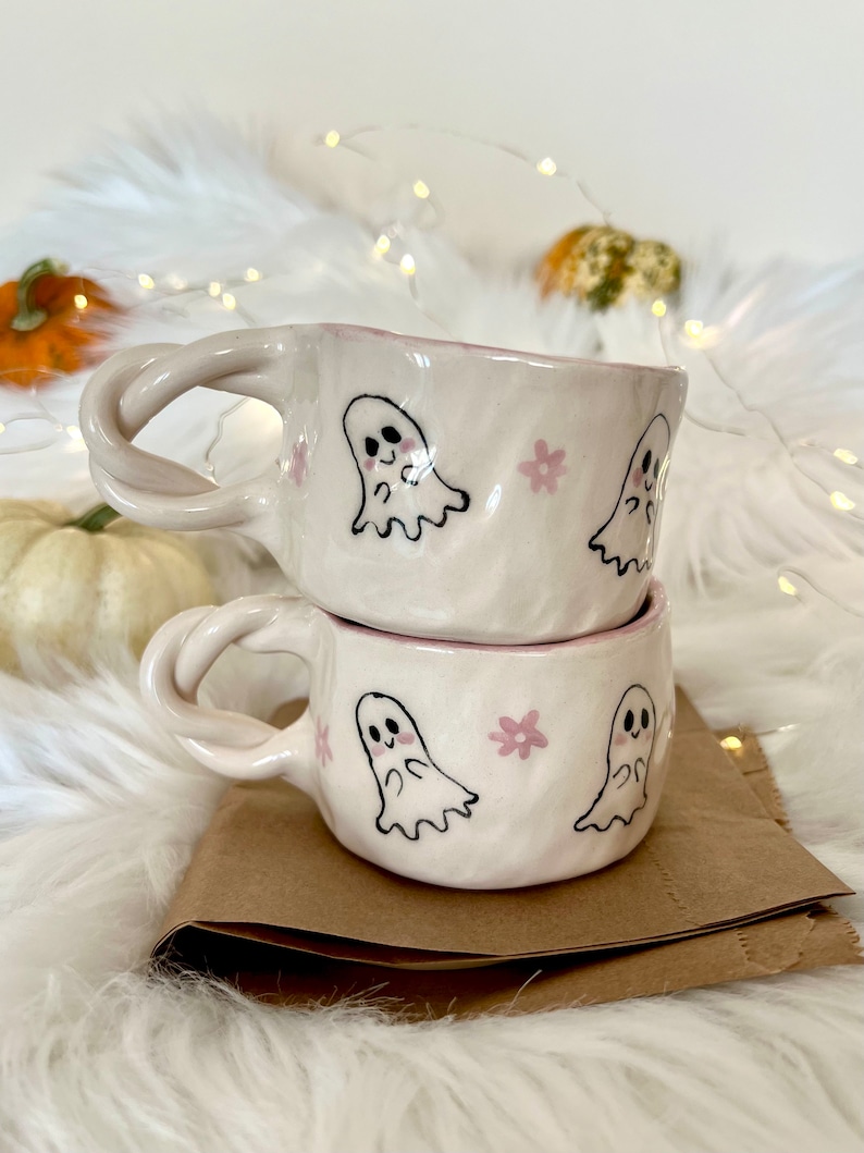 May include: Two white ceramic mugs with pink and black ghost illustrations and pink flowers. The mugs are stacked on top of each other.