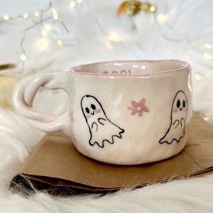 May include: White ceramic mug with a pink rim and a pink floral design. The mug features two hand-drawn ghosts with black eyes and pink cheeks.