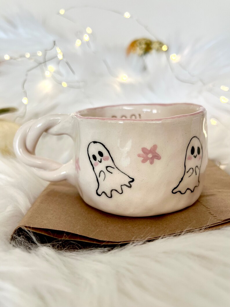May include: White ceramic mug with a pink rim and a pink floral design. The mug features two hand-drawn ghosts with black eyes and pink cheeks.