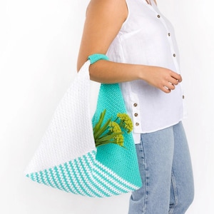 May include: A handmade, triangular crochet tote bag. The bag features a white section and a turquoise section with white stripes. The bag is filled with green plants and flowers. The handle is turquoise.