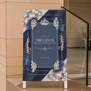 Puede incluir: Un letrero de cumplea&ntilde;os azul marino y blanco con el texto "Bienvenida al 50 cumplea&ntilde;os de Miranda" y la fecha "07.25.20XX". El letrero est&aacute; decorado con perlas, cintas y una corona.