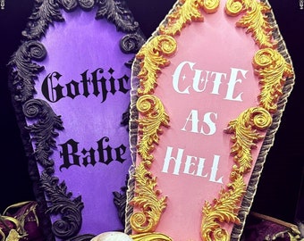 May include: Two decorative coffin-shaped signs with ornate gold and black trim. The purple sign says "Gothic Babe" and the pink sign says "Cute as Hell".