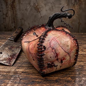 May include: A pumpkin-shaped object with a pale, textured surface featuring red vein-like markings and dark splatters. The segments are joined by thick, black, visible stitching. A dark, twisted stem sits on top, and a metal cleaver lies nearby.