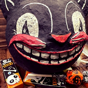 May include: A black paper mache cat mask with a wide, toothy grin and red painted lips. The mask is surrounded by candy sticks and lollipops with skull and crossbones designs.