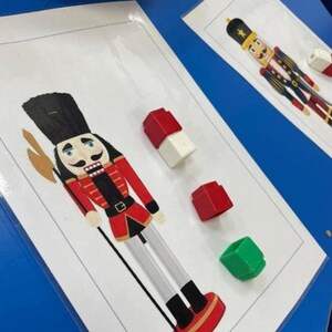 Könnte beinhalten: Ein rotes, weißes und schwarzes Nussknacker-Spielzeug mit einem roten und schwarzen Hut und einer roten und schwarzen Uniform. Das Spielzeug hält einen goldenen Stab. Neben dem Spielzeug befinden sich vier bunte Blöcke.