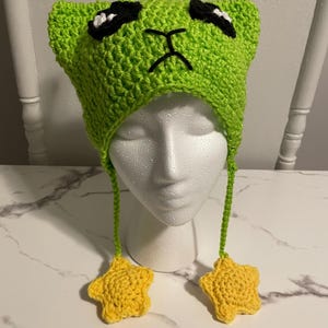 Puede incluir: Sombrero de extraterrestre de color verde tejido a crochet con antenas en forma de estrella amarilla y lazos colgantes en forma de estrella amarilla. El sombrero tiene ojos negros y una boca fruncida.