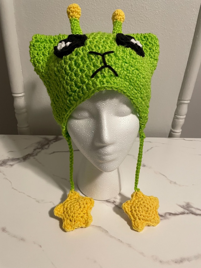 Puede incluir: Sombrero de extraterrestre de color verde tejido a crochet con antenas en forma de estrella amarilla y lazos colgantes en forma de estrella amarilla. El sombrero tiene ojos negros y una boca fruncida.
