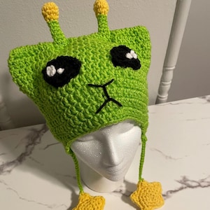 Puede incluir: Sombrero de extraterrestre de color verde tejido a crochet con ojos negros, nariz blanca y antenas en forma de estrella amarilla. El sombrero tiene una larga correa verde con extremos en forma de estrella amarilla.