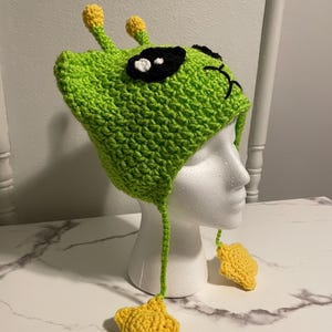 Puede incluir: Sombrero de extraterrestre de crochet verde con ojos negros y adornos de estrellas amarillas.