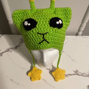 Puede incluir: Sombrero de extraterrestre de color verde tejido a crochet con antenas en forma de estrella amarilla y lazos colgantes en forma de estrella amarilla. El sombrero tiene ojos negros y una boca negra.