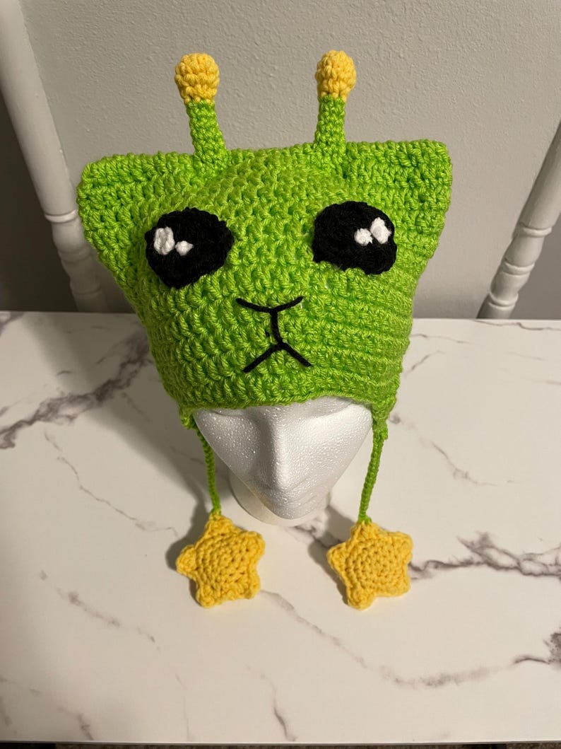 Puede incluir: Sombrero de extraterrestre de color verde tejido a crochet con antenas en forma de estrella amarilla y lazos colgantes en forma de estrella amarilla. El sombrero tiene ojos negros y una boca negra.