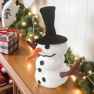 May include: A handmade snowman decoration made of white yarn with a black top hat, coal buttons, and a carrot nose. The snowman has brown twig arms and a stitched smile. Festive holiday decor.