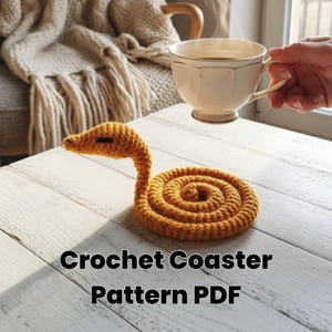 May include: A crocheted orange snake-shaped coaster sits on a white wooden table. The coaster is coiled with a raised head. The text "Crochet Coaster Pattern PDF" is at the bottom of the image. A teacup is held in the background.