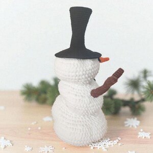 May include: A crocheted snowman with a black top hat, an orange carrot nose, and a brown arm. The snowman is white with a textured surface and is decorated with snowflakes. The snowman is a winter holiday decoration.