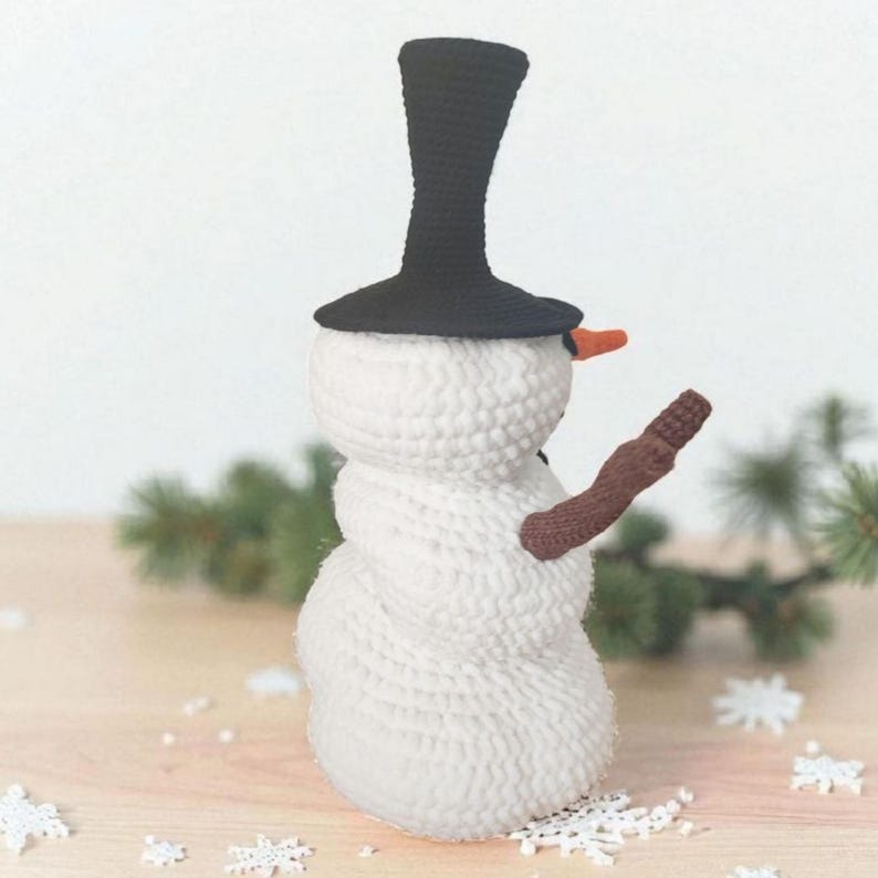 May include: A crocheted snowman with a black top hat, an orange carrot nose, and a brown arm. The snowman is white with a textured surface and is decorated with snowflakes. The snowman is a winter holiday decoration.