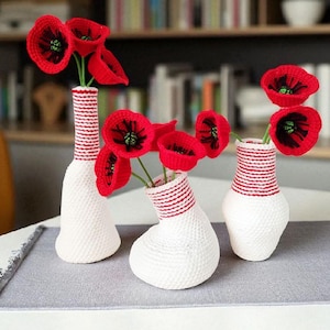 Puede incluir: Tres jarrones blancos de ganchillo con bordes a rayas rojas y blancas, cada uno con amapolas rojas de ganchillo vibrantes con centros negros. Los jarrones están dispuestos sobre una superficie gris, mostrando una decoración floral hecha a mano.