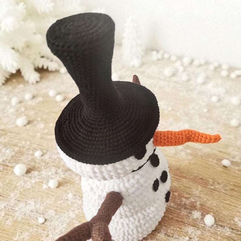 May include: A handmade crochet snowman with a black top hat, orange carrot nose, and brown twig arms. The snowman has black button eyes and a white body. The winter-themed decoration is set against a snowy background.