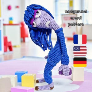 May include: A handmade amigurumi doll in shades of blue, purple, and tan, with the text "amigurumi ennui pattern." The doll has long blue hair and is posed in a slumped position. Flags of France, the United States, and Germany are also visible.