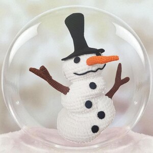 May include: A crocheted snowman with a black top hat, orange carrot nose, and brown twig arms is encased in a clear glass globe. The snowman has black button eyes and a stitched mouth. The snowman is a winter holiday decoration.