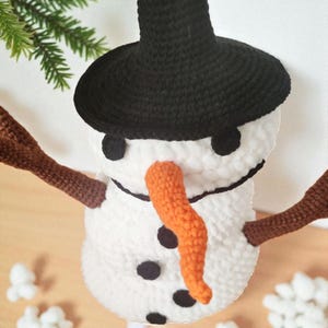 May include: A handmade crochet snowman ornament with a black hat, eyes, and buttons. It has a bright orange carrot nose and brown stick arms. The snowman is white and fluffy, perfect for winter holiday decor.