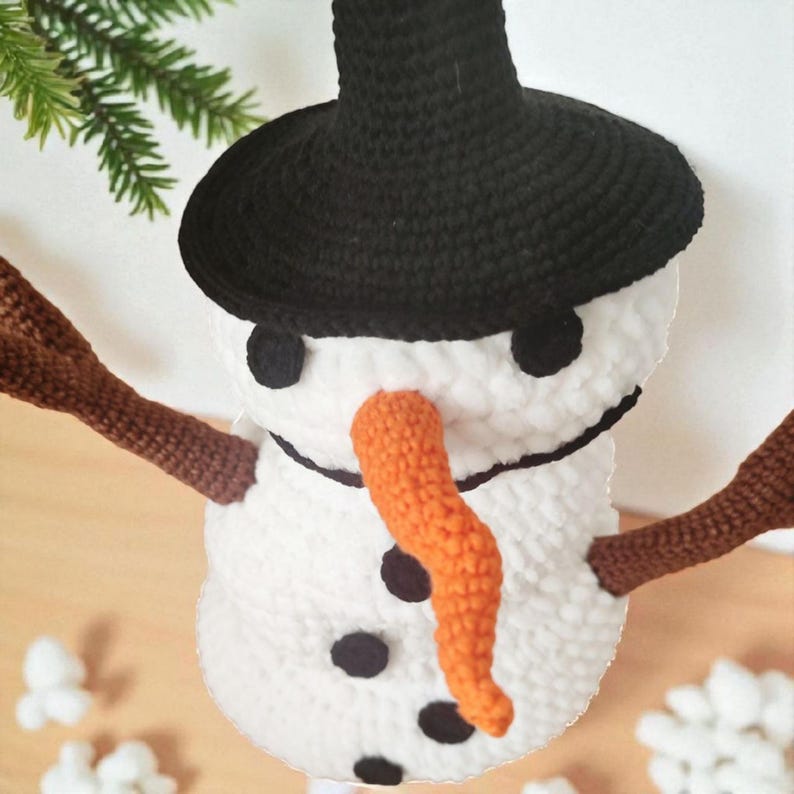 May include: A handmade crochet snowman ornament with a black hat, eyes, and buttons. It has a bright orange carrot nose and brown stick arms. The snowman is white and fluffy, perfect for winter holiday decor.