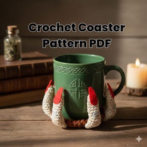 May include: A green ceramic mug with a Celtic knot design, resting on a crocheted coaster shaped like claws with red felt accents. The image includes the text "Crochet Coaster Pattern PDF". A candle and a small jar are in the background.