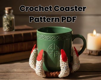 May include: A green ceramic mug with a Celtic knot design, resting on a crocheted coaster shaped like claws with red felt accents. The image includes the text "Crochet Coaster Pattern PDF". A candle and a small jar are in the background.