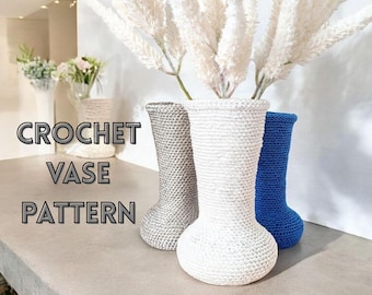 May include: Three crochet vases in white, silver, and blue, each holding fluffy white pampas grass. The vases have a bulbous base and a narrow neck. Text on the left reads "CROCHET VASE PATTERN".