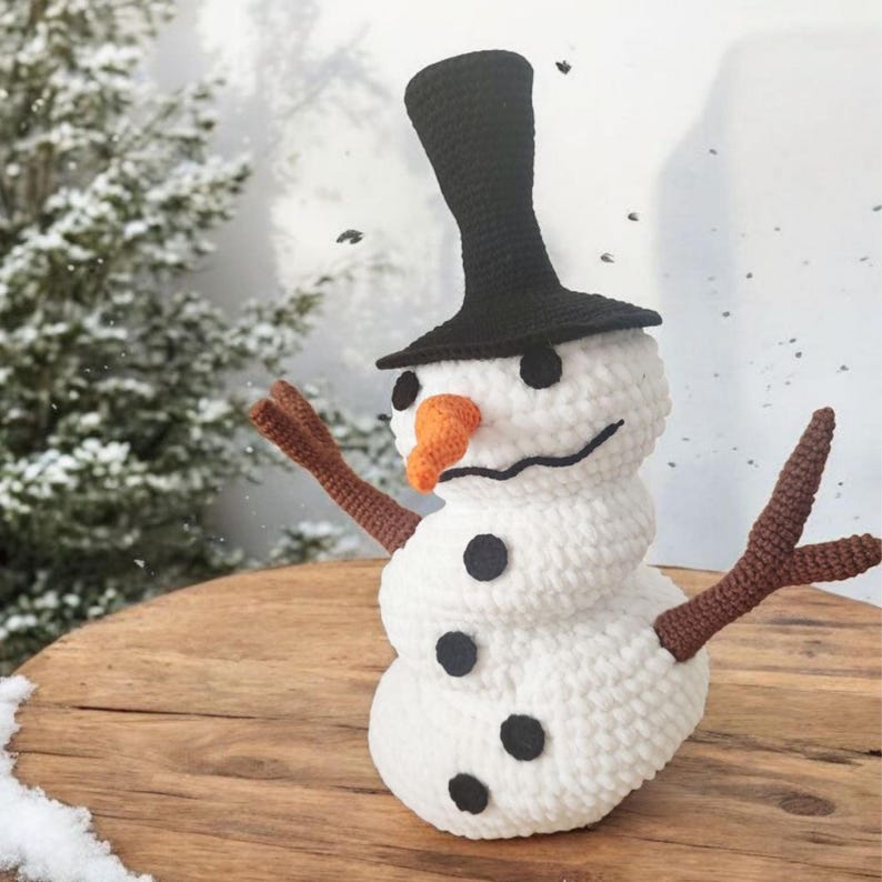 May include: A handmade crochet snowman with a black top hat, black button eyes, and an orange carrot nose. The snowman has brown stick arms and black button accents. It is a winter-themed decoration.