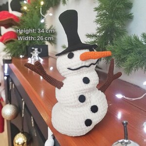 May include: A crocheted snowman with a black top hat, orange carrot nose, and brown twig arms. The snowman is white with black button eyes and a black mouth. Height 13.4 inches, width 10.2 inches. Festive holiday decor.