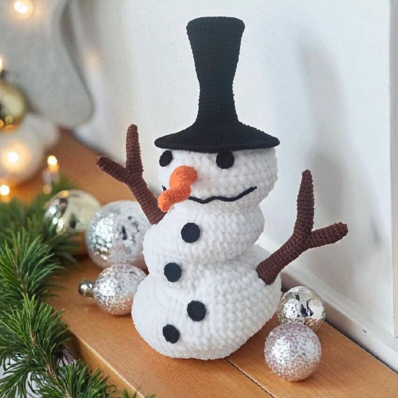 May include: A handmade crochet snowman with a black top hat, orange carrot nose, and twig arms. The snowman has black button eyes and a stitched smile. Decorative silver ornaments and pine branches are in the background.