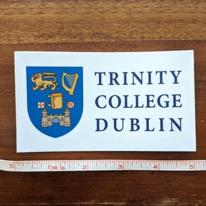 May include: A white rectangular sticker with the words "TRINITY COLLEGE DUBLIN" in navy blue. The sticker features a blue crest with a gold lion, harp, and other symbols. A measuring tape is placed below the sticker.