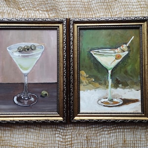K&ouml;nnte beinhalten: Zwei gerahmte Gem&auml;lde von Martini-Gl&auml;sern mit Oliven. Die Gl&auml;ser sind mit einer klaren Fl&uuml;ssigkeit gef&uuml;llt und mit einer gr&uuml;nen Olive garniert. Die Gem&auml;lde sind in goldenen Rahmen und haben einen wei&szlig;en Hintergrund.