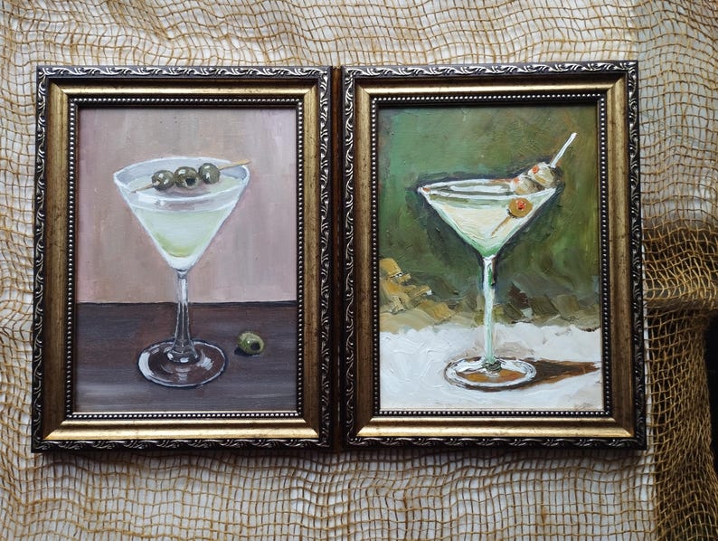 K&ouml;nnte beinhalten: Zwei gerahmte Gem&auml;lde von Martini-Gl&auml;sern mit Oliven. Die Gl&auml;ser sind mit einer klaren Fl&uuml;ssigkeit gef&uuml;llt und mit einer gr&uuml;nen Olive garniert. Die Gem&auml;lde sind in goldenen Rahmen und haben einen wei&szlig;en Hintergrund.