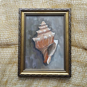 Può includere: Dipinto a olio di una conchiglia in una cornice dorata decorativa. La conchiglia è dipinta in tonalità pesca, marrone e bianco su uno sfondo grigio. La cornice ha un design ornato. L'opera d'arte è esposta su un tessuto beige strutturato.