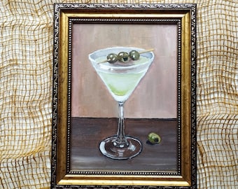 May include: An oil painting in a decorative gold frame depicting a martini glass filled with a pale green liquid. Three green olives are skewered on a toothpick resting across the rim, and one single green olive sits on the dark brown surface.