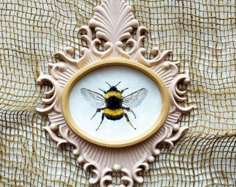 May include: A framed oval artwork featuring a detailed illustration of a bumblebee. The bee has black and yellow stripes and translucent wings. The frame is ornate, in a soft pink color, with a gold-colored inner border.