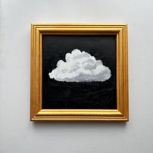 May include: A framed painting featuring a white cloud on a black canvas. The cloud has a textured, fluffy appearance. The gold-coloured rectangular frame provides a classic aesthetic to the artwork.