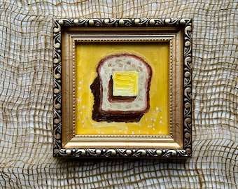 Puede incluir: Pintura al óleo enmarcada de una rebanada de pan tostado con mantequilla. La pintura tiene un fondo amarillo y un marco dorado adornado. La tostada es marrón y blanca.